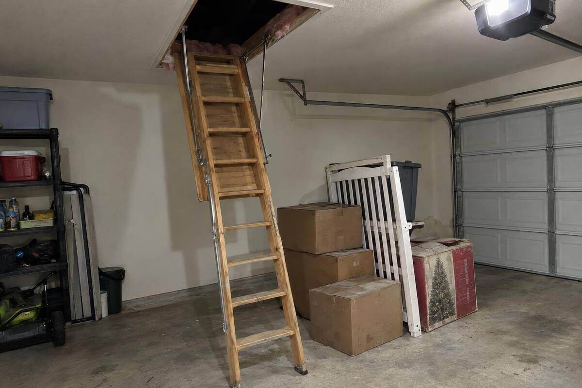 Dusty cardboard boxes and a folded crib stacked at the foot of a pull-down attic ladder in a garage