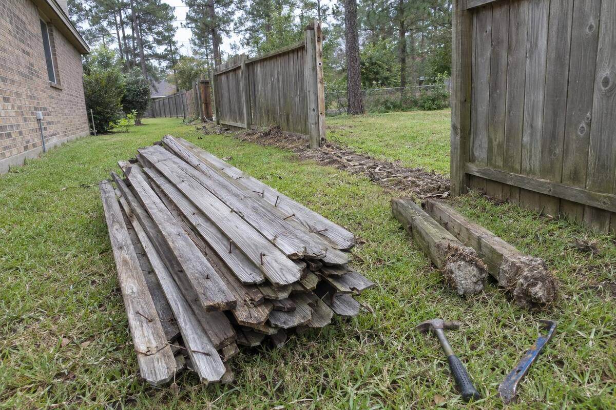 Gray weathered fence pickets and rails stacked along a side yard with bent nails still in them