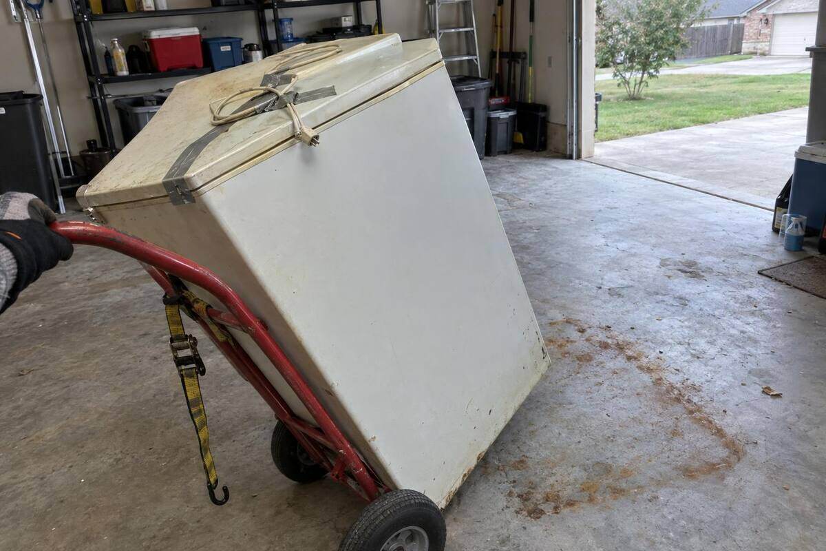 An old white chest freezer tipped back on a hand truck inside a garage, its cord taped to the lid