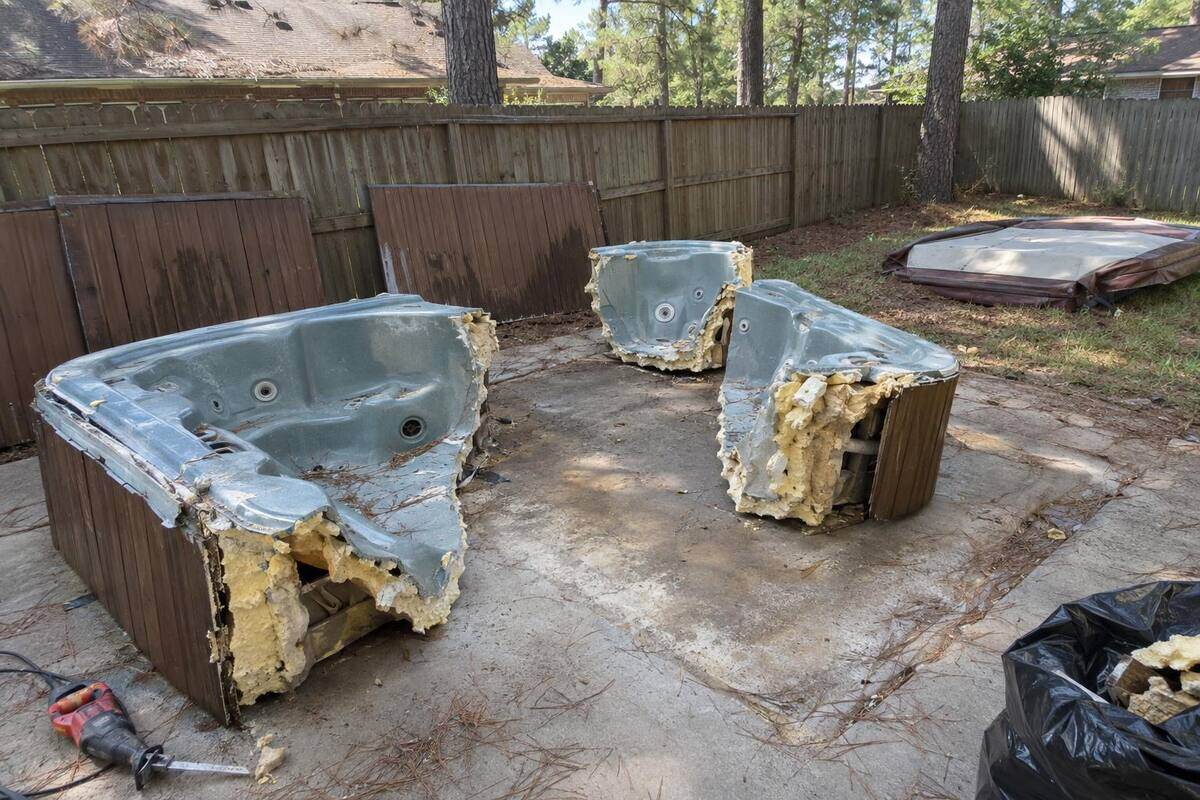 An old hot tub shell cut into sections on a backyard patio, its wood skirting panels leaning on a fence