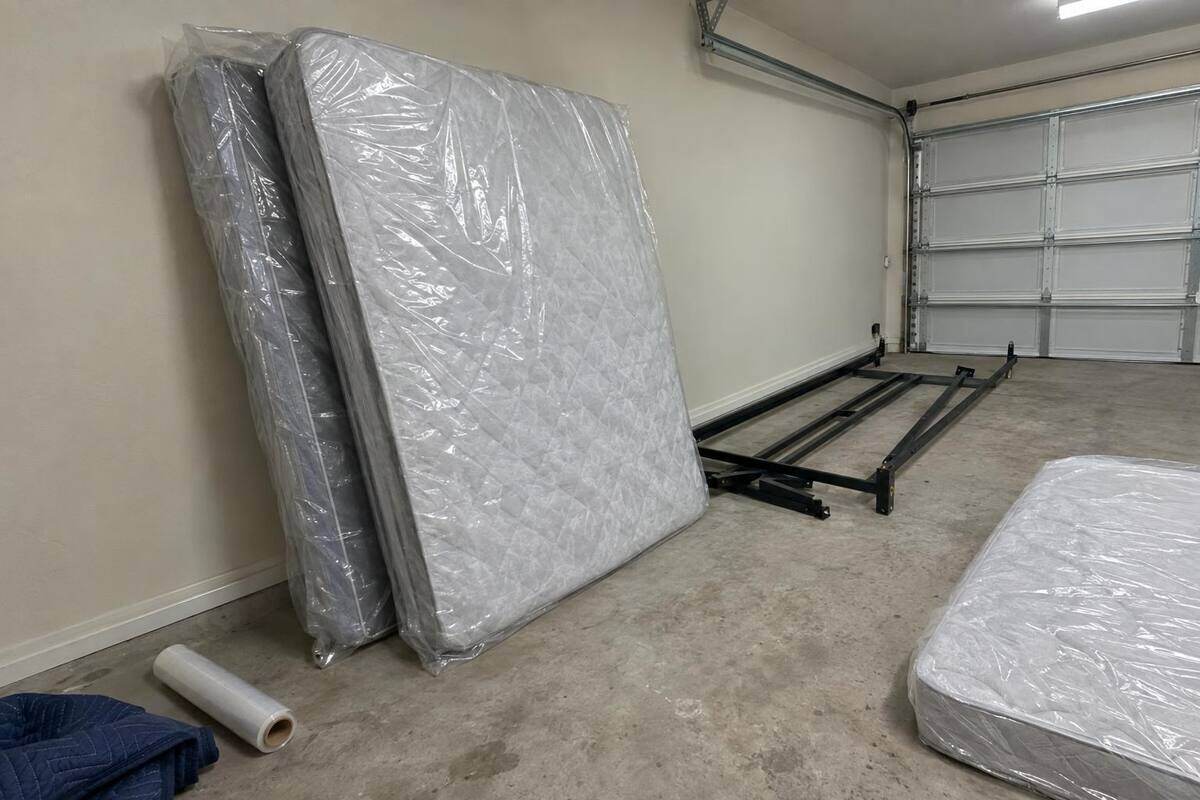 A mattress and box spring wrapped in clear plastic, leaning against a garage wall