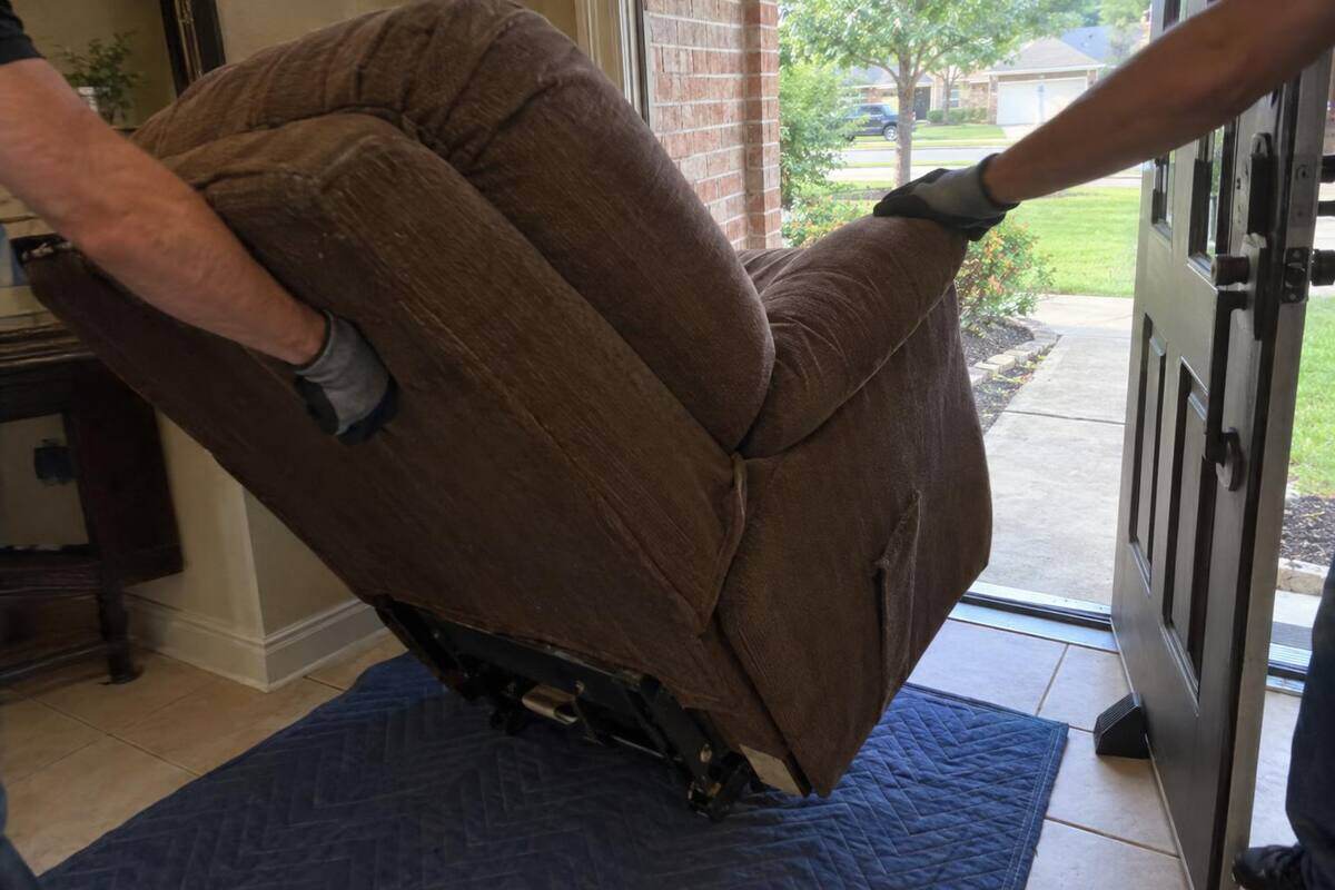 Gloved hands tilting a worn recliner through a front doorway, the door propped open